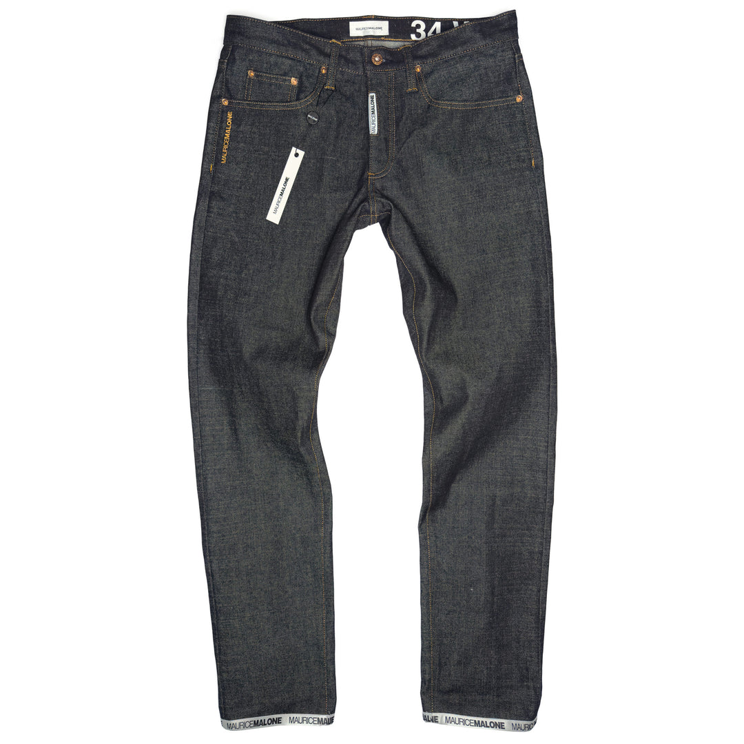 A pair of Maurice Malone jeans in raw 15 oz. Japanese selvedge denim, featuring a woven logo cuff, branded fly labeling, copper rivets, and orange stitching. The jeans are displayed flat, showcasing their handcrafted details and modern slim fit.